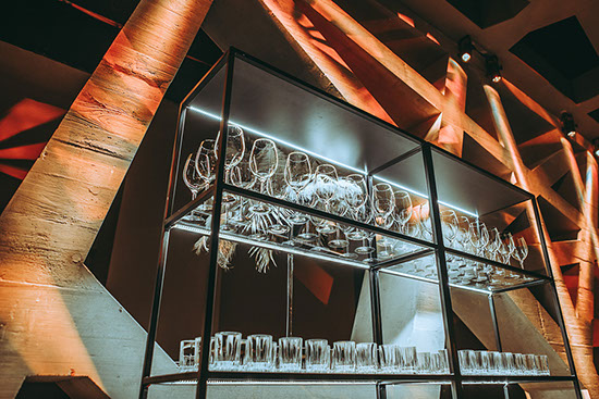bar-rueckwaende_palais-berg - Palais Wertheim Wien Industrial glassware rack with upside-down wine glasses and tumblers on two shelves, lit by warm bar lighting in a modern setting.