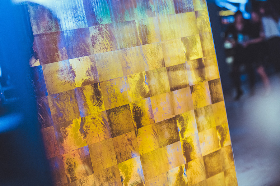 deko-waende_palais-berg - Palais Wertheim Wien Close-up of a gold-tiled mosaic wall reflecting yellow and gold hues, with blue lighting on the left and a blurred crowd in the background.