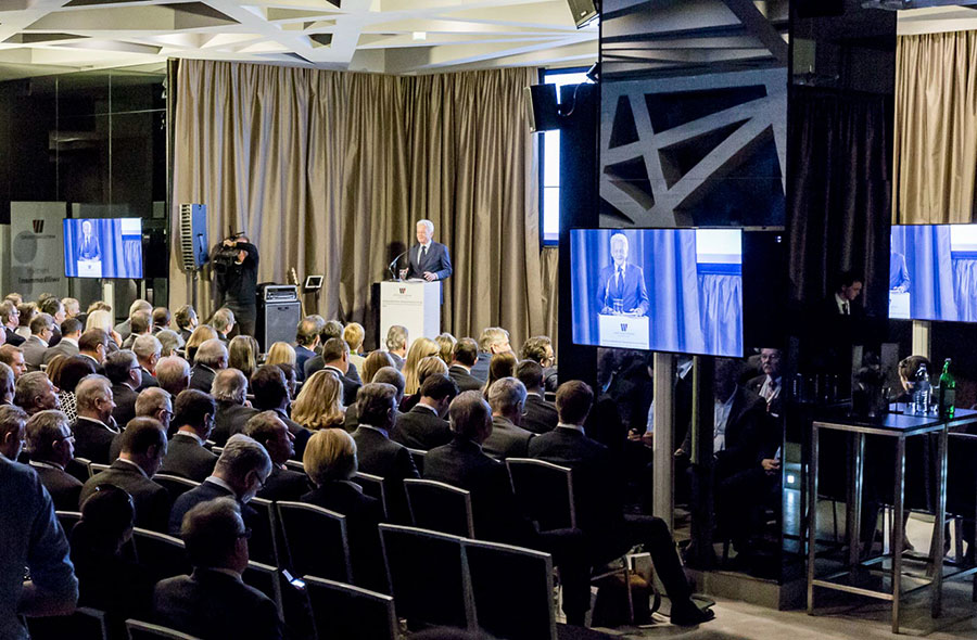 flatscreen-55_palais-berg - Palais Wertheim Wien Speaker at a podium addressing a large seated audience in a conference room with video screens displaying the speaker nearby