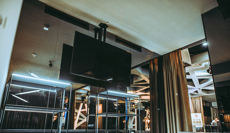 flatscreen-75_palais-berg - Palais Wertheim Wien Large flat-screen TV mounted above glass-and-metal shelving in a modern lobby with blue neon lighting.