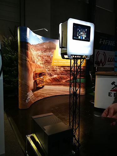 palais-wertheim-fotobox-1 - Palais Wertheim Wien Touchscreen display mounted on a tall stand at an expo booth, with a desert quarry backdrop and a yellow truck image behind it.