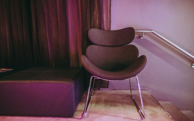 Modern curved-back lounge chair with chrome legs in a purple-lit corner beside a purple padded bench and metal railing.