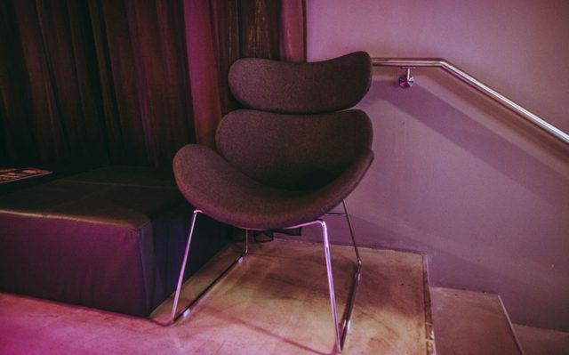 Modern brown upholstered lounge chair with metal legs, placed in a dim purple-lit corner near a staircase rail.