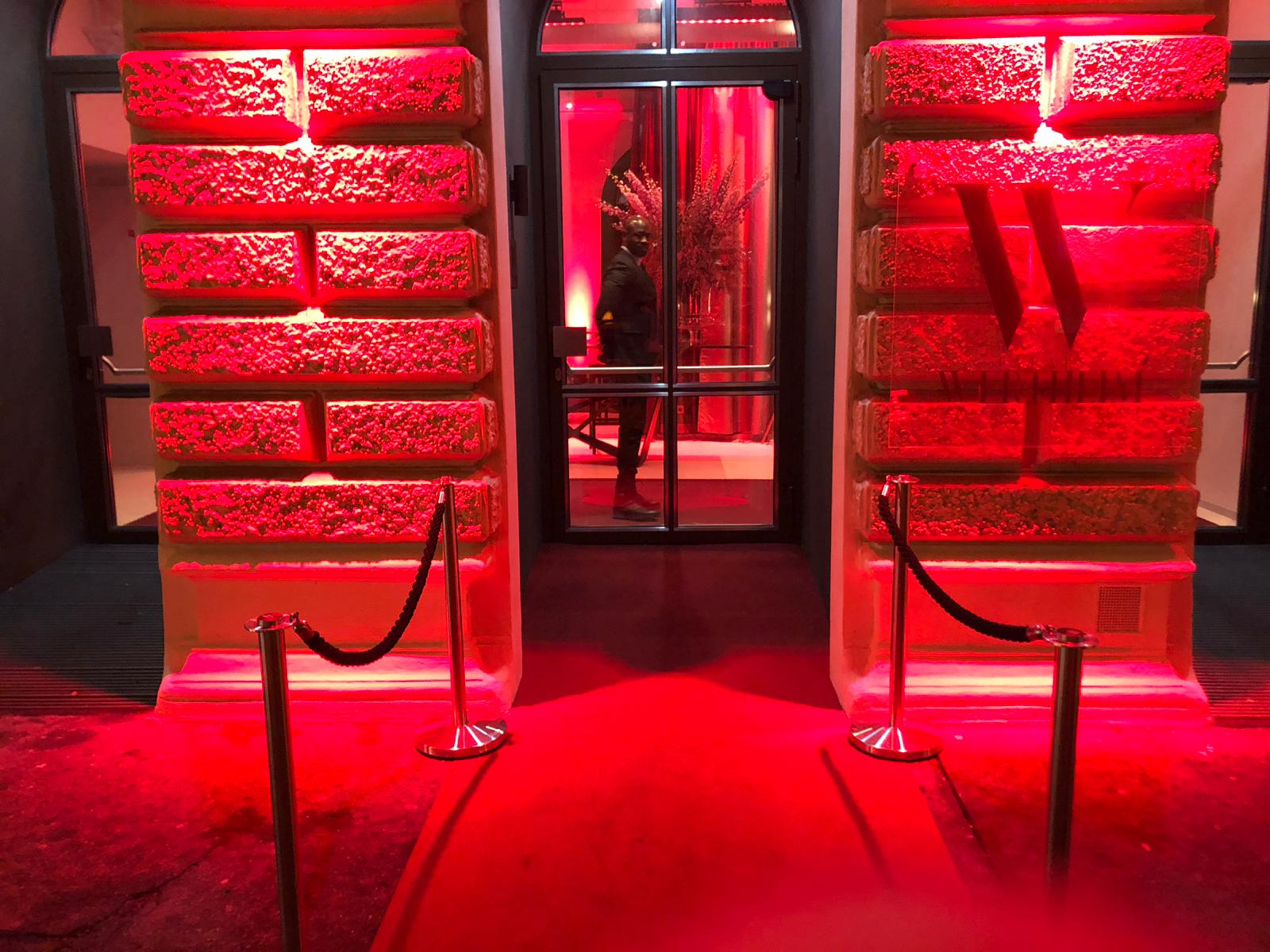 palais-wertheim_lichtanlage_foyer-fassade - Palais Wertheim Wien Red-lit entrance framed by textured stone columns, glass doors in the middle, with rope barriers on either side and a person inside near the door.",