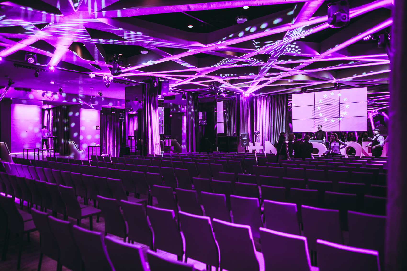 palais-wertheim_lichtanlage_wertheim-saal - Palais Wertheim Wien Conference hall with purple lighting, empty rows of chairs, and a stage with a large screen displaying a circular EU-like emblem.