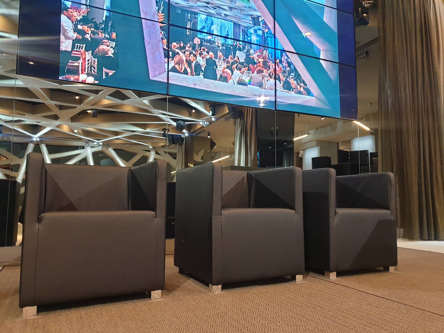 podium-sessel_palais-wertheim-2 - Palais Wertheim Wien Four black armchairs in a modern lounge facing a large multi-screen video wall showing a crowded event.