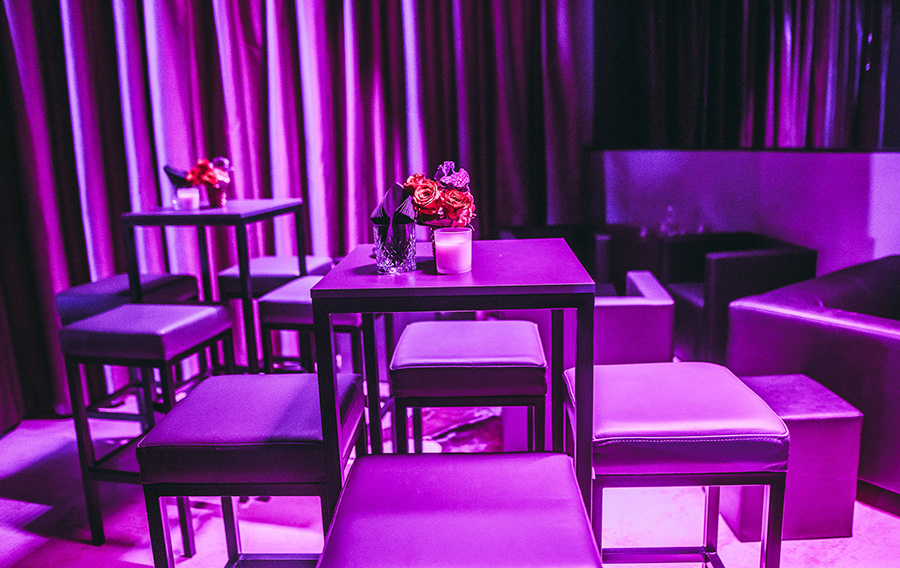 stehpule_50x_palais-berg - Palais Wertheim Wien Dimly lit lounge with square tables, purple lighting, and cushioned stools; small flower arrangements and candles on tables.