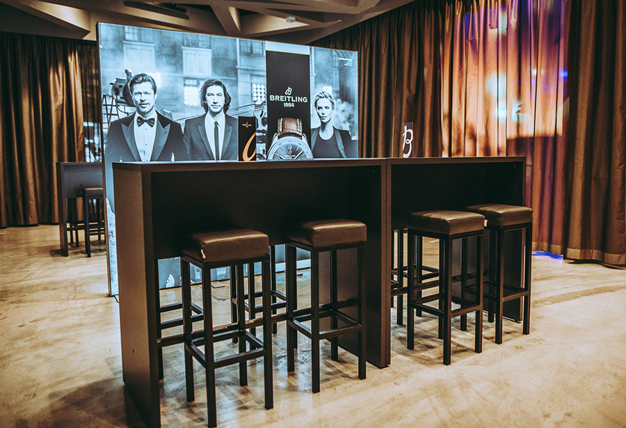 stehpule_palais-berg - Palais Wertheim Wien Breitling promotional booth with black high tables and stools, backdrop featuring brand imagery and watches in a curtained event space