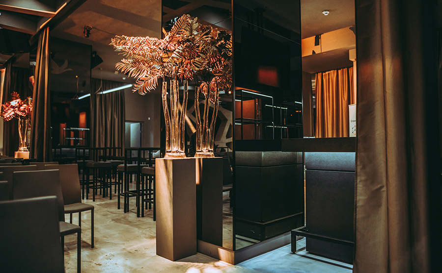 vasenpodeste_palais-berg - Palais Wertheim Wien Upscale lounge interior with warm lighting, tall glass vases with ferns on pedestals, and dark seating.